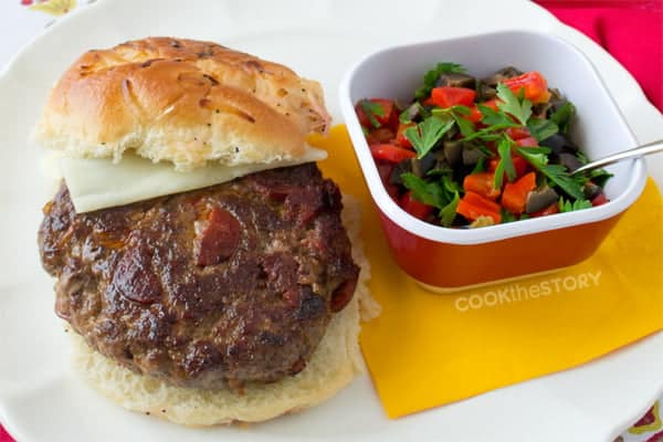 Flamenco burger with cheese on a bun, dish of chopped olives, peppers, and parsley to the side.