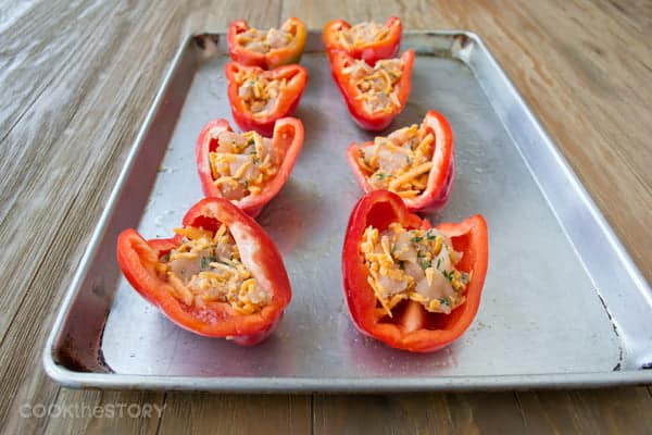 Halved red bell peppers with raw chicken and cheese inside on a baking sheet.