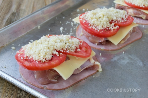 Ham, chicken, slice of cheese, tomato slices, and breadcrumbs in stacks on a baking sheet, not yet baked.