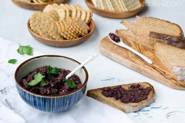 Bowl of tapanade, tapenade spread on slice of bread, with more bread and crackers in background.
