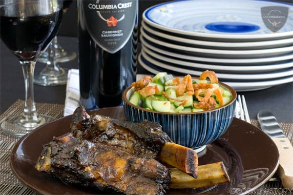 Beef Ribs on a brown plate with a blue bowl of cucumber salad, red wine in the background.