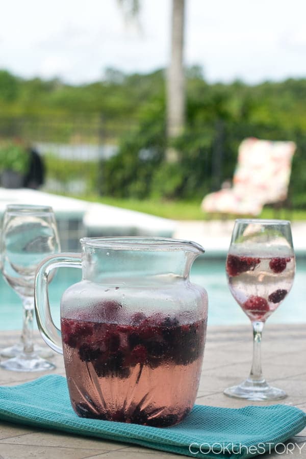 Glass pitcher and wine glass filled with Berry Wine Cooler by a pool.