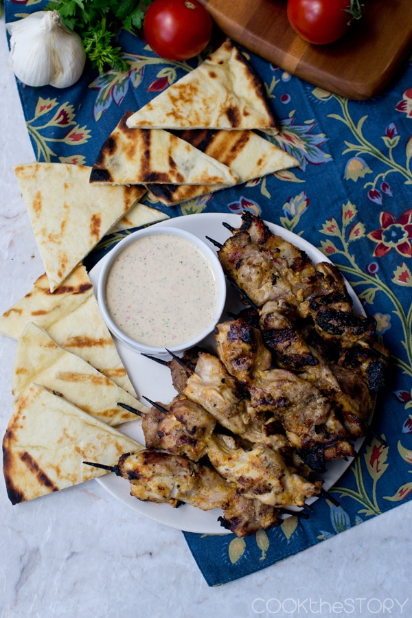 Overhead view of plate with grilled chicken kebabs, yogurt sauce, and grilled pita wedges.