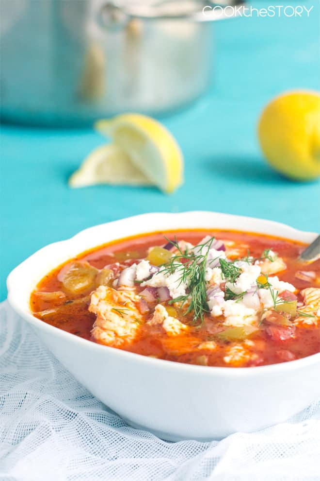 Greek Seafood Soup with a tomato broth and topped with Greek salad ingredients, in a white bowl.
